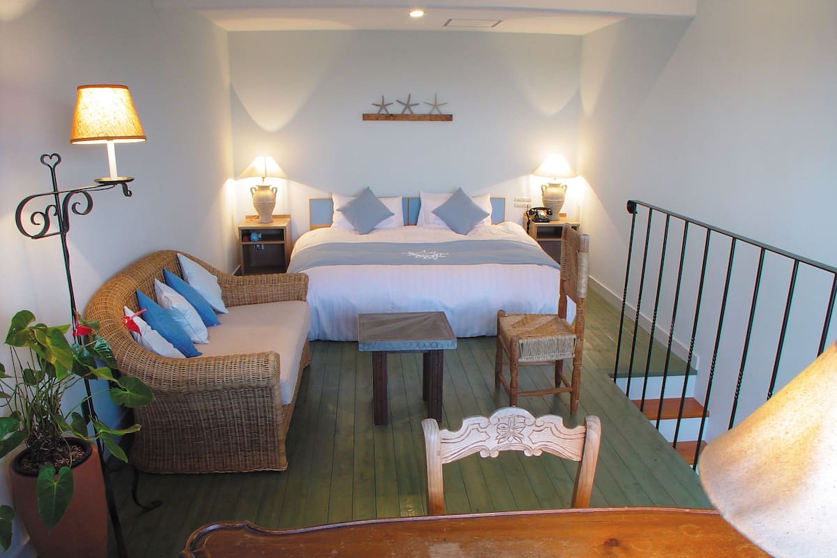A bedroom with a bed, two bedside tables with lamps, a wicker chair, a wooden chair, and a potted plant.
