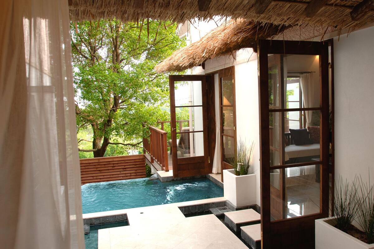 A private pool area with a wooden deck, a glass door leading to a seating area, and a thatched roof structure.