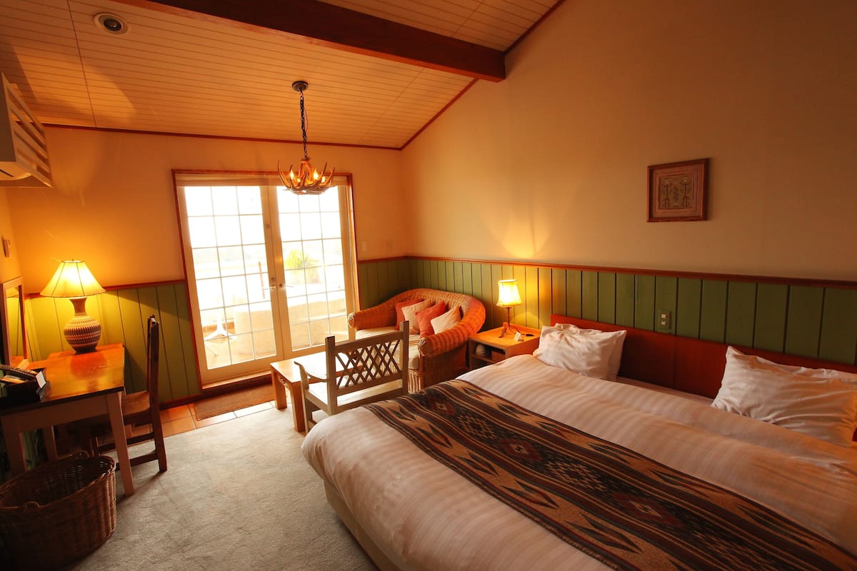A hotel room with a large bed, a desk with a lamp, a chair, and a view of the outside through a sliding door.