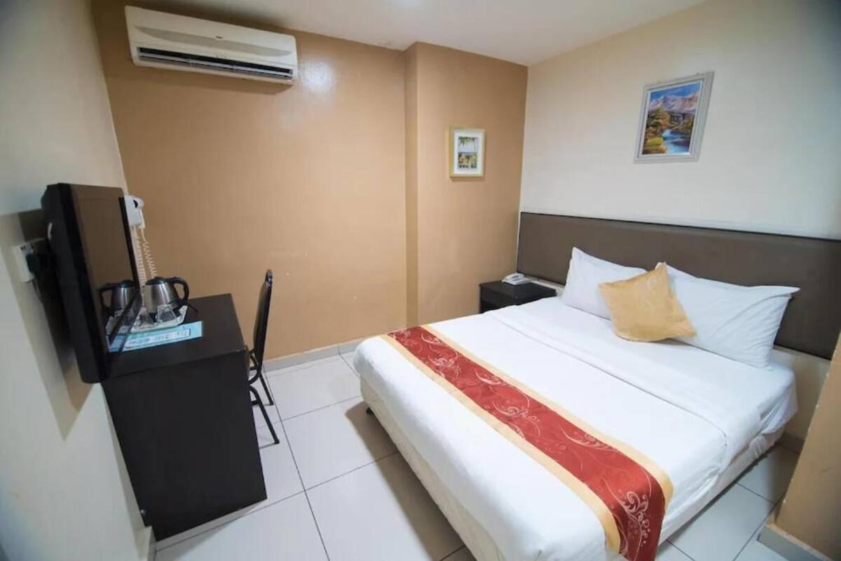 A hotel room with a bed, a nightstand, a desk with a telephone, and an air conditioner.