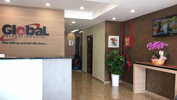 Uma área de recepção de hotel com balcão, uma grande planta em vaso e uma TV fixada na parede.