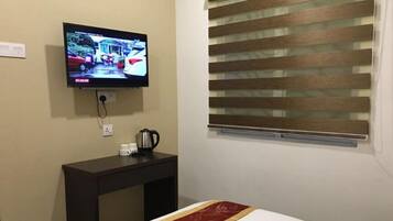 A hotel room with a bed, a TV mounted on the wall, a small table with a kettle and cups, and a window with blinds.