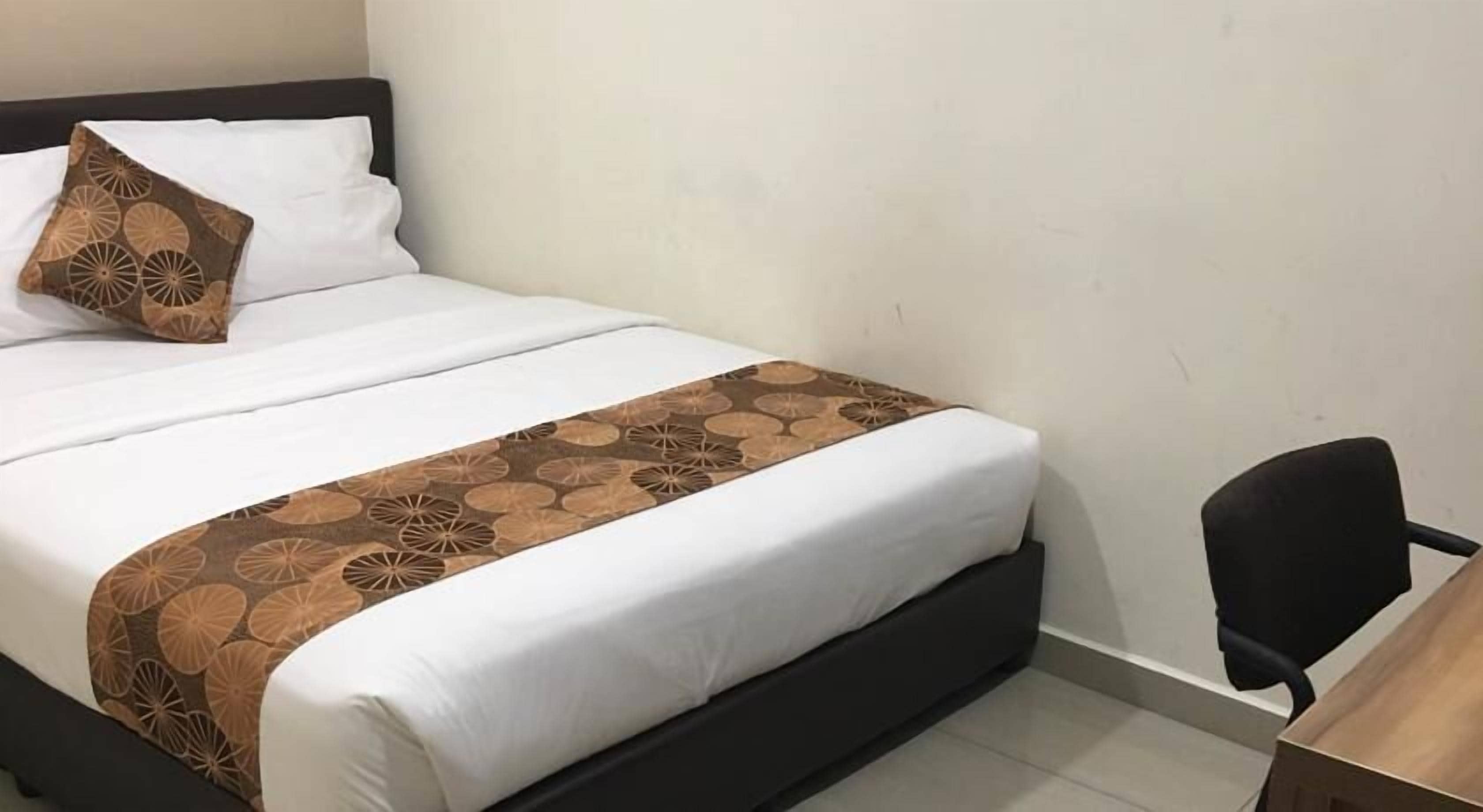 A neatly made bed with a patterned bedspread and pillows, a black chair, and a wooden desk in a simple room with plain walls.