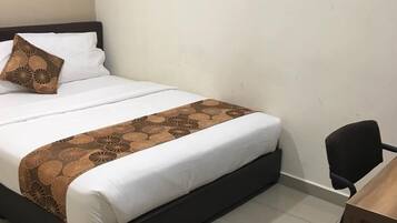 A neatly made bed with a patterned bedspread and pillows, a black chair, and a wooden desk in a simple room with plain walls.
