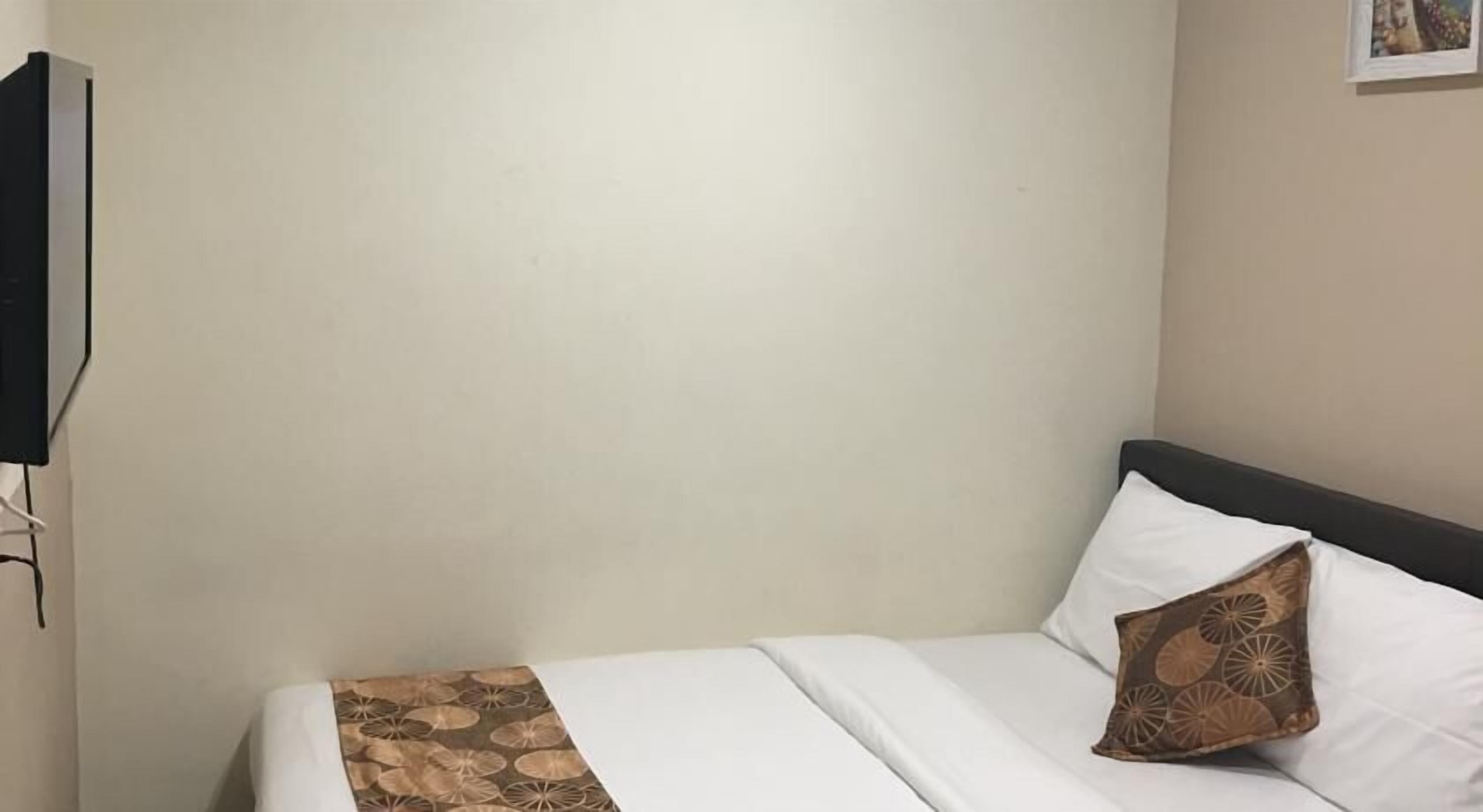 A neatly made bed with a white sheet and a patterned throw blanket, a single brown pillow, and a plain wall with a framed picture.