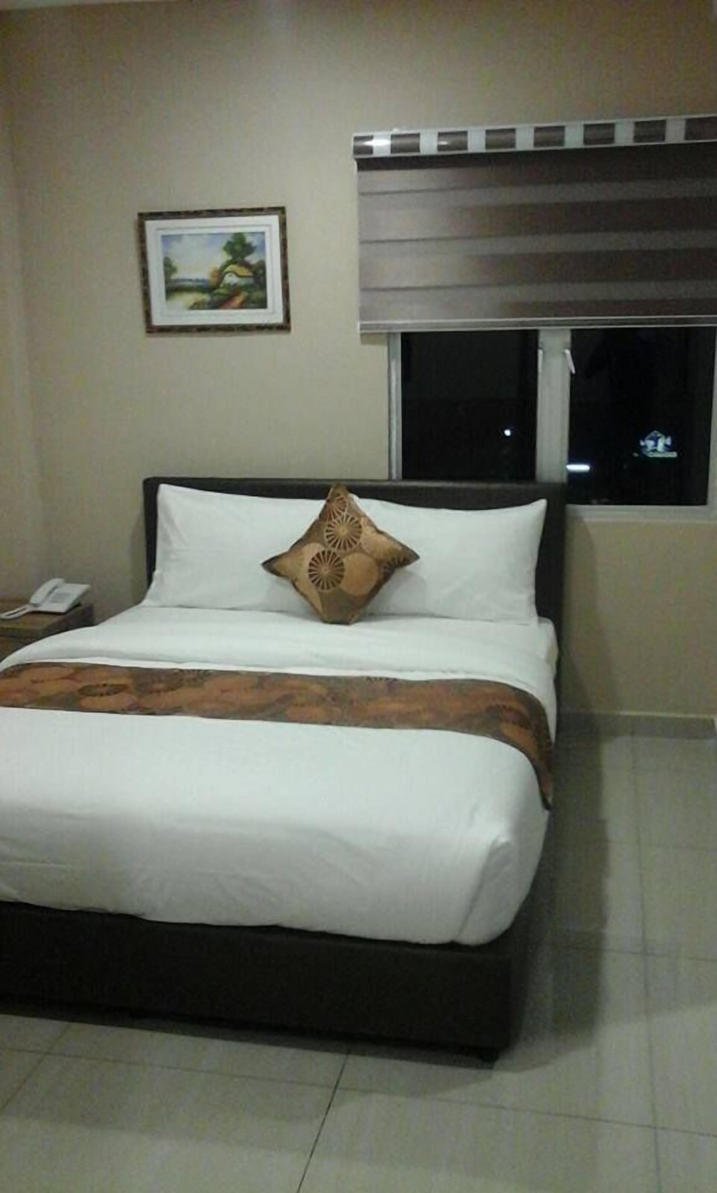 A hotel room with a bed, a nightstand with a phone, a window with blinds, and a framed picture on the wall.