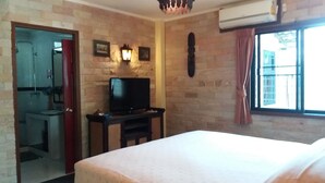 A hotel room with a bed, a television on a wooden stand, a stone wall, a window with curtains, and a bathroom visible through an open door. - Baan Laksee Pool Villa (Pattaya)