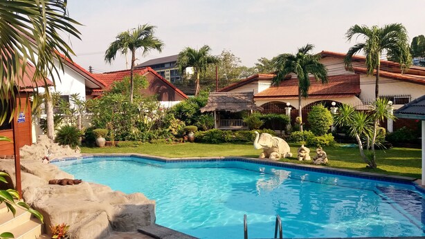 A swimming pool surrounded by lush greenery and palm trees, with a stone sculpture and a thatched-roof structure nearby. - Baan Laksee Pool Villa (Pattaya)