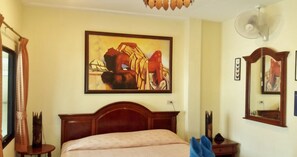 A hotel room with a bed, a painting on the wall, a fan, and a mirror.