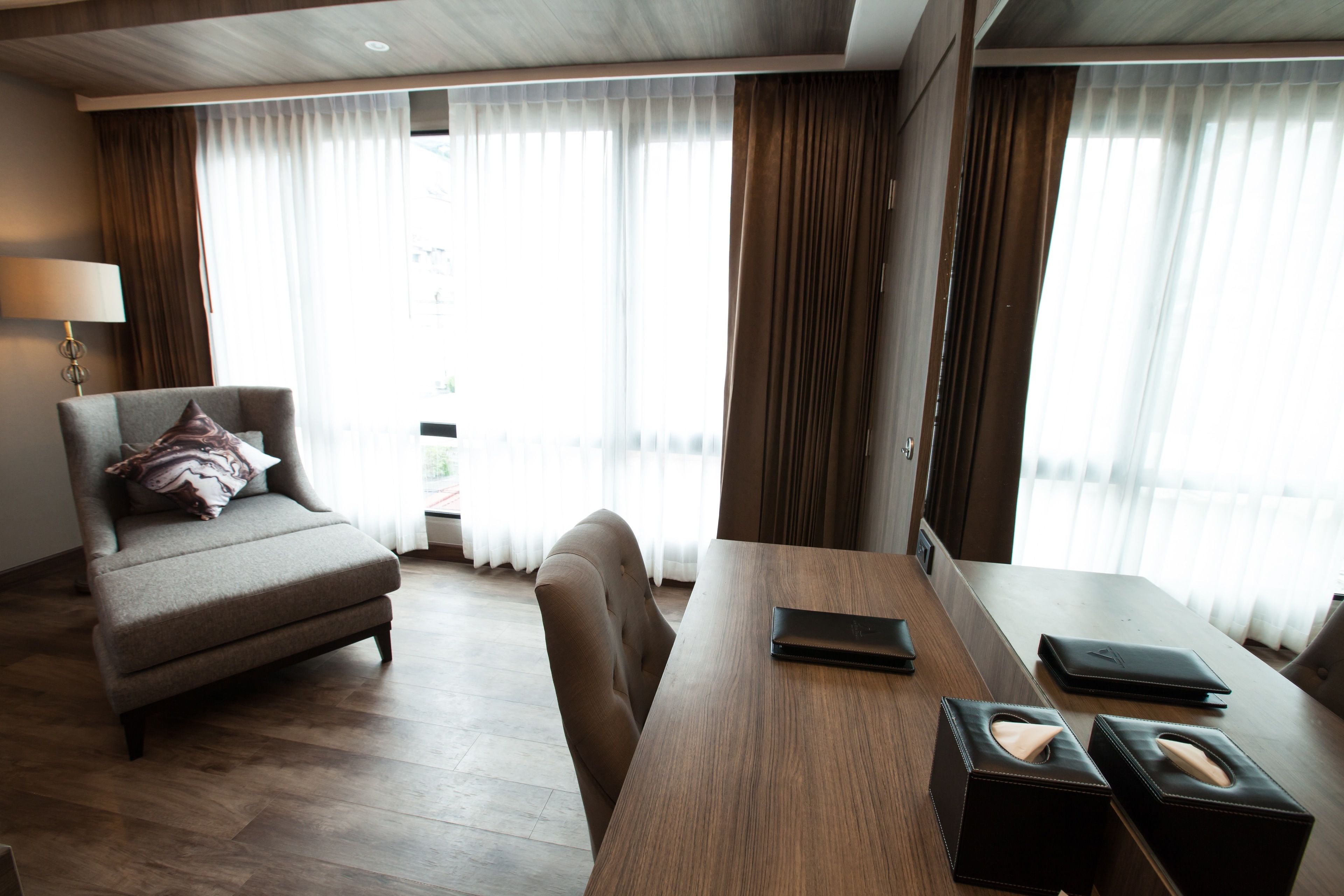 A modern hotel room with a sofa, a wooden dining table, and a view through large windows with curtains.