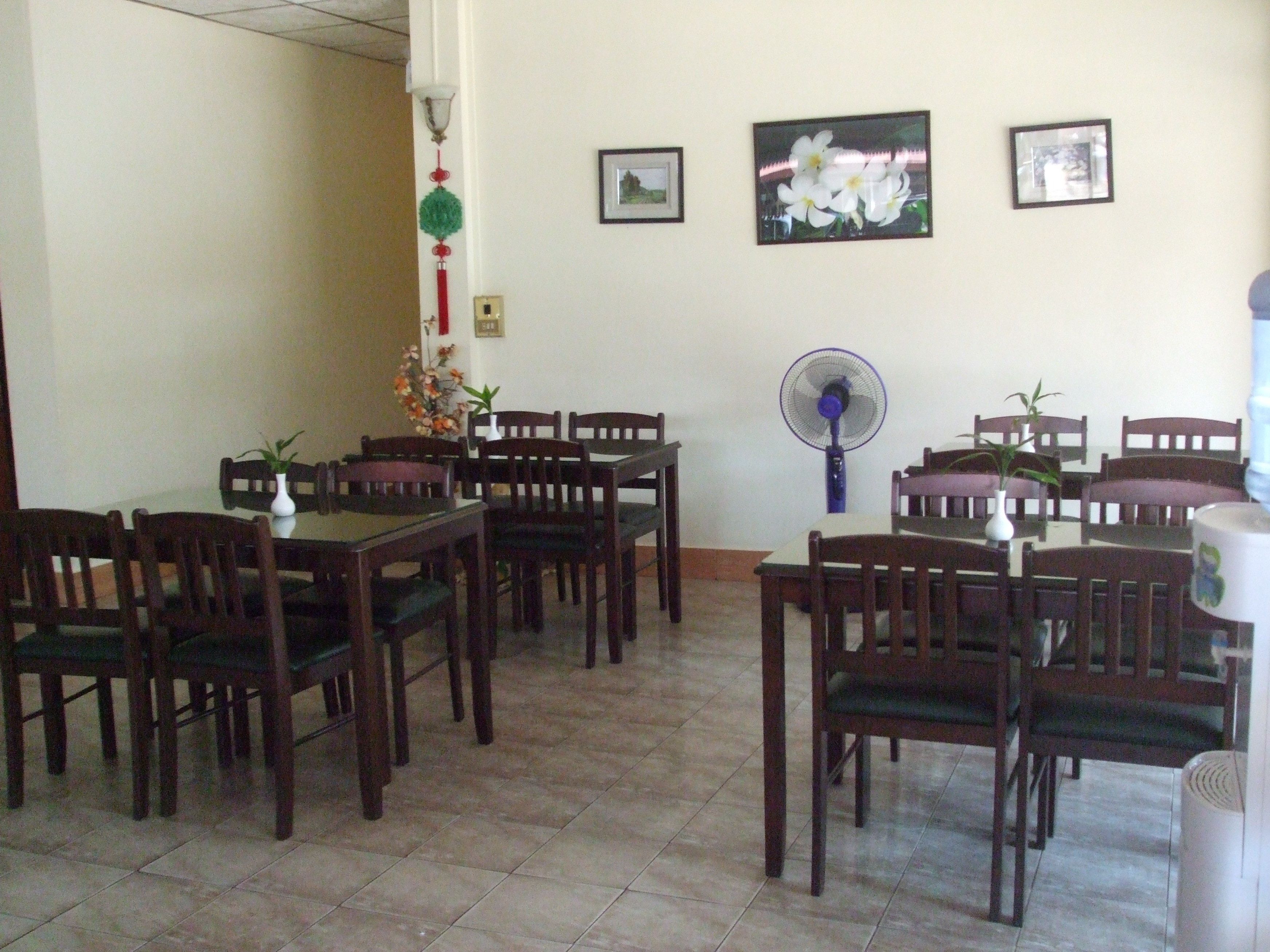 Un comedor con mesas y sillas de madera, un ventilador de pie y plantas decorativas.