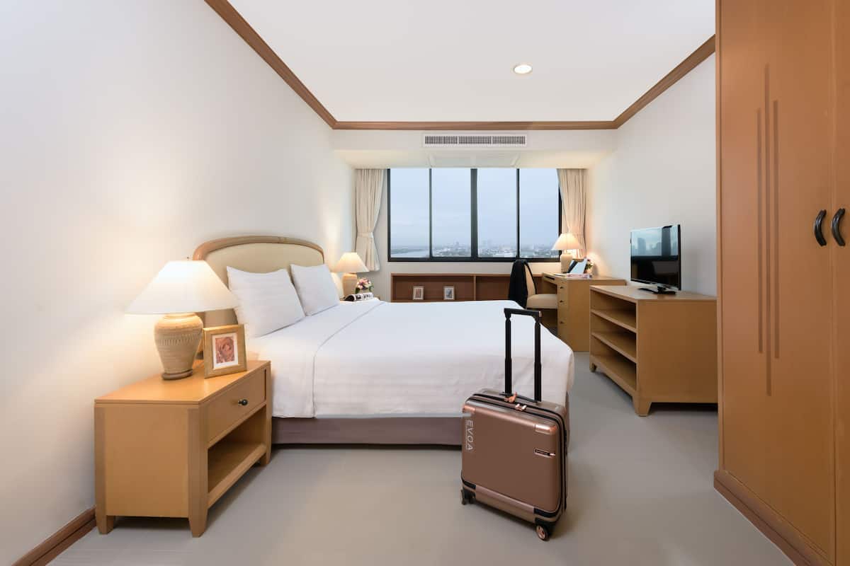Une chambre d’hôtel avec un lit, des lampes de chevet, un bureau avec une télévision, une valise et une grande fenêtre donnant sur la ville.