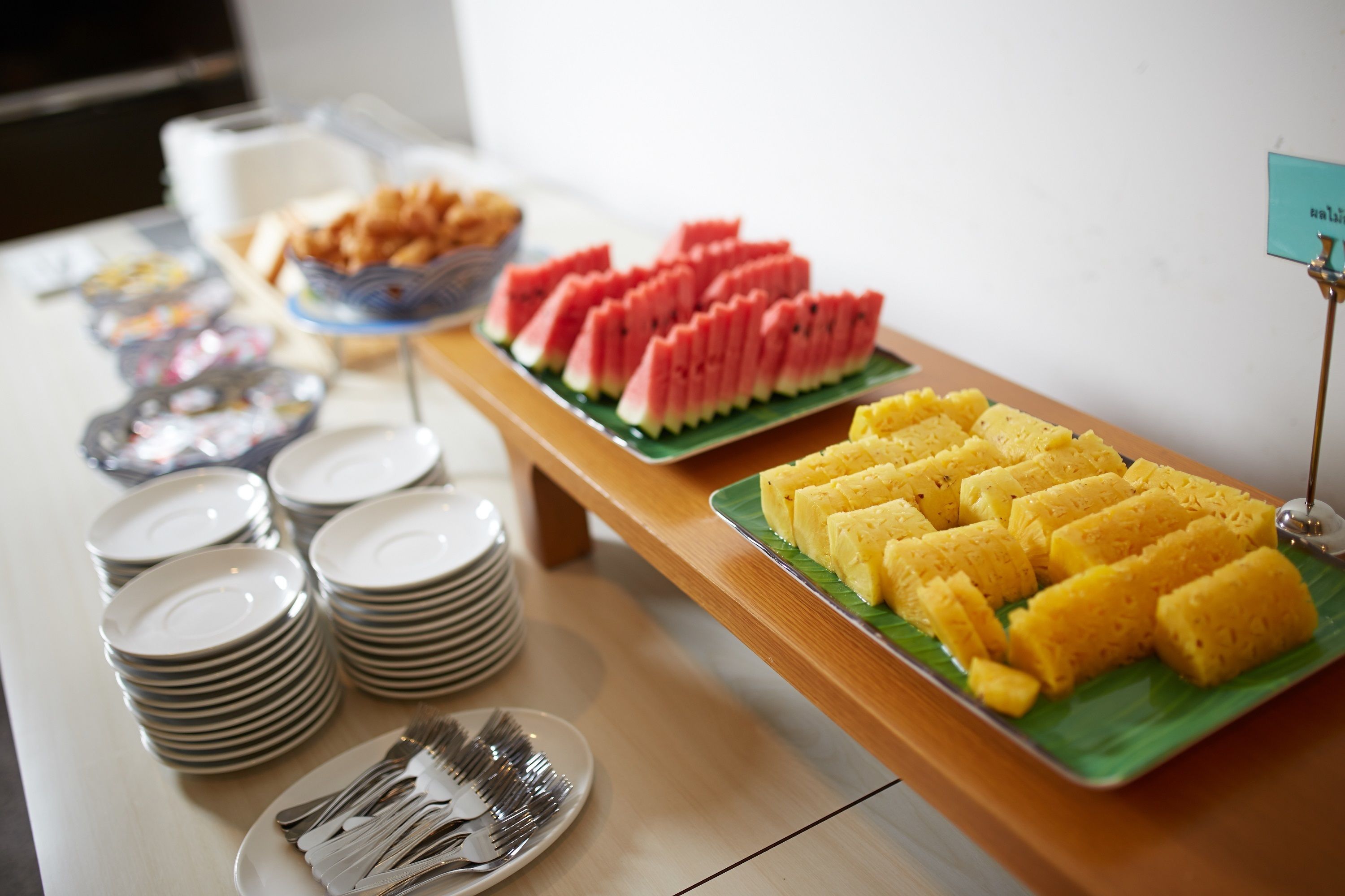 A buffet table with plates of food, including watermelon and a yellow dish, and stacks of white plates and forks.