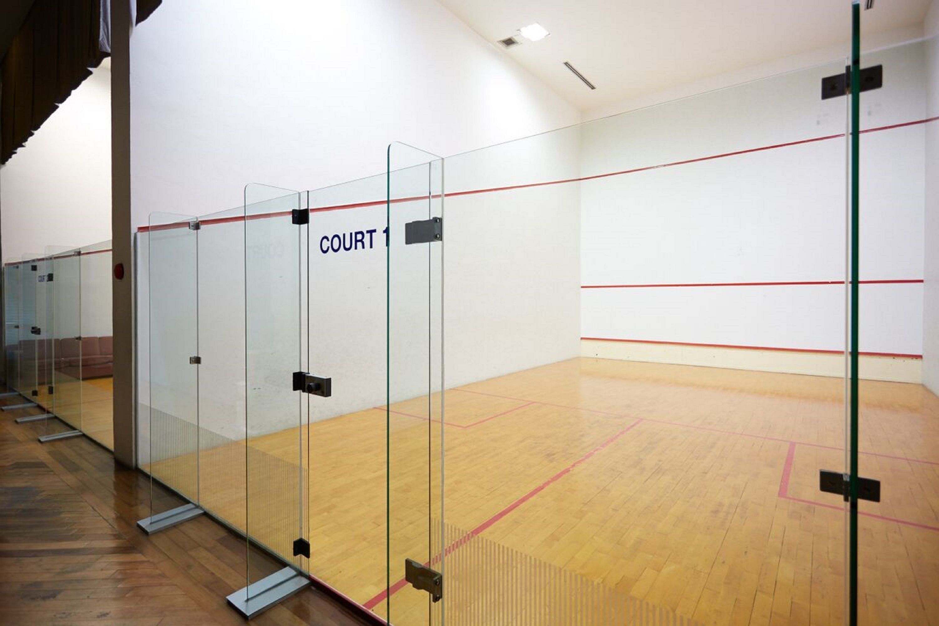 A squash court with glass walls, wooden floor, and a red line marking the playing area.