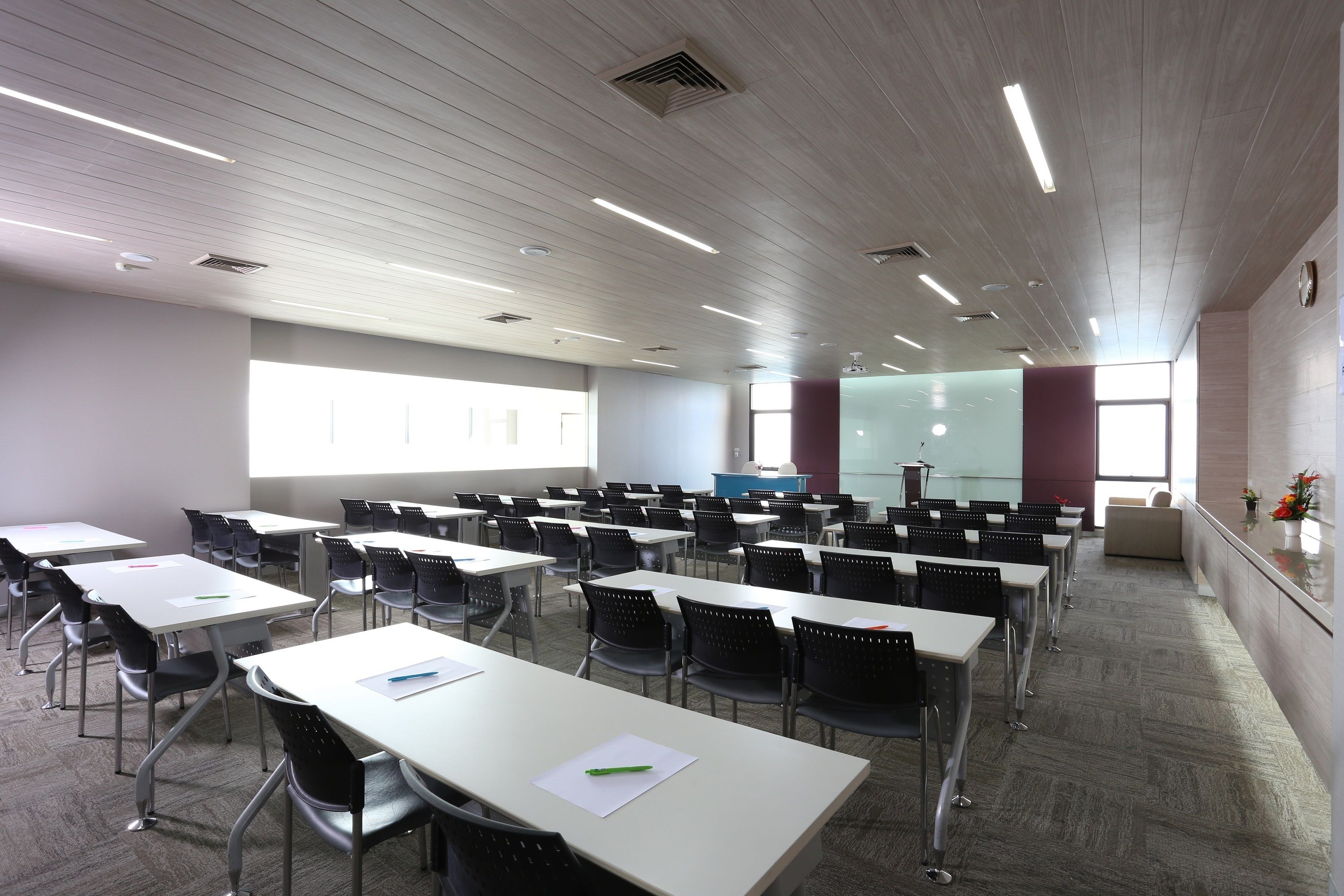 A classroom with rows of tables and chairs, a projector screen, and a window providing natural light.