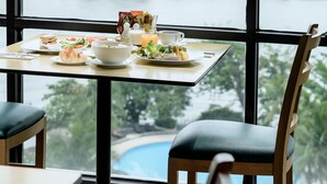 Una mesa de comedor con comida y bebidas, una silla, y vistas a un río y edificios a través de amplios ventanales.