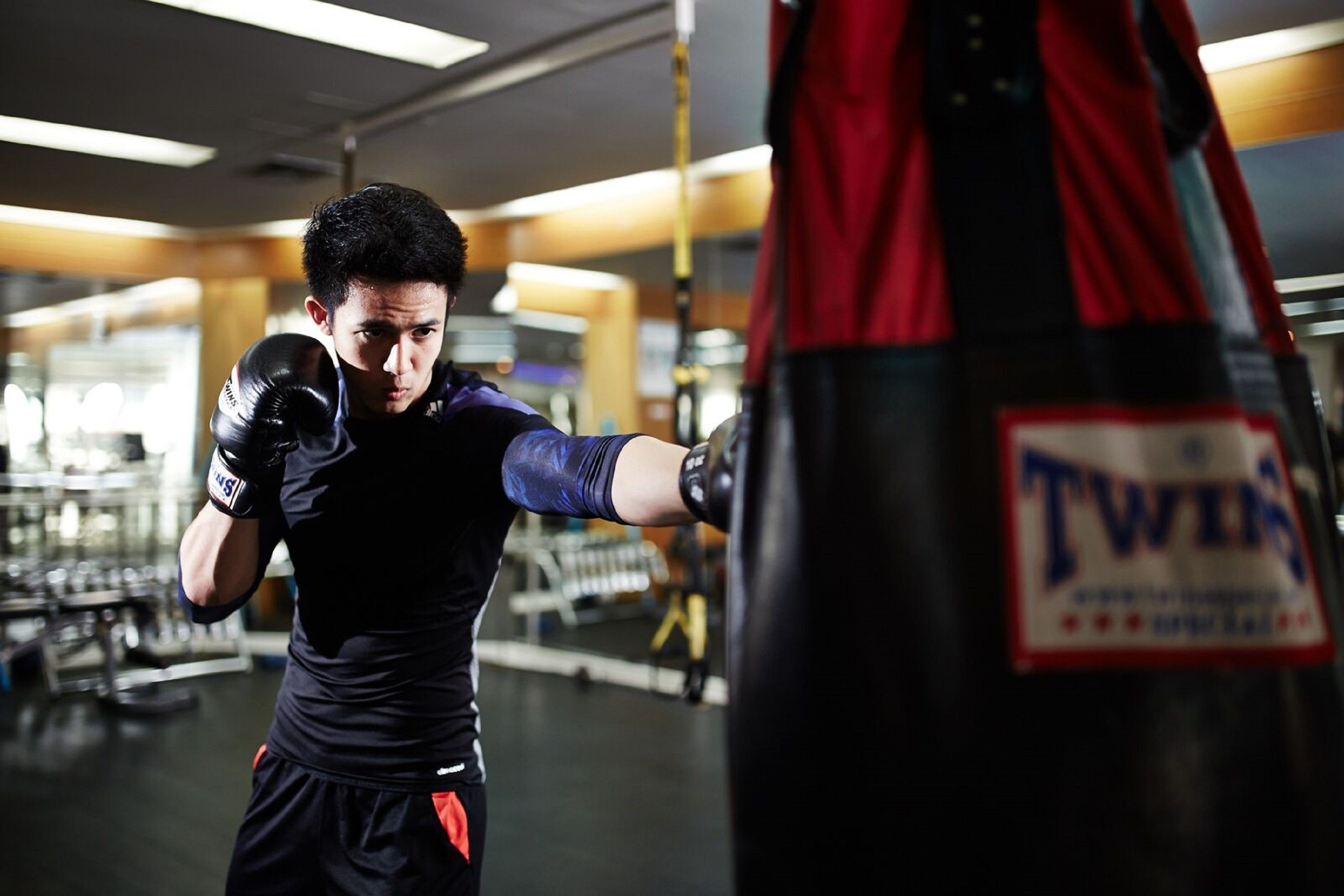 A person practicing boxing with a punching bag labeled "Twins" in a gym with visible equipment and a mirrored wall.