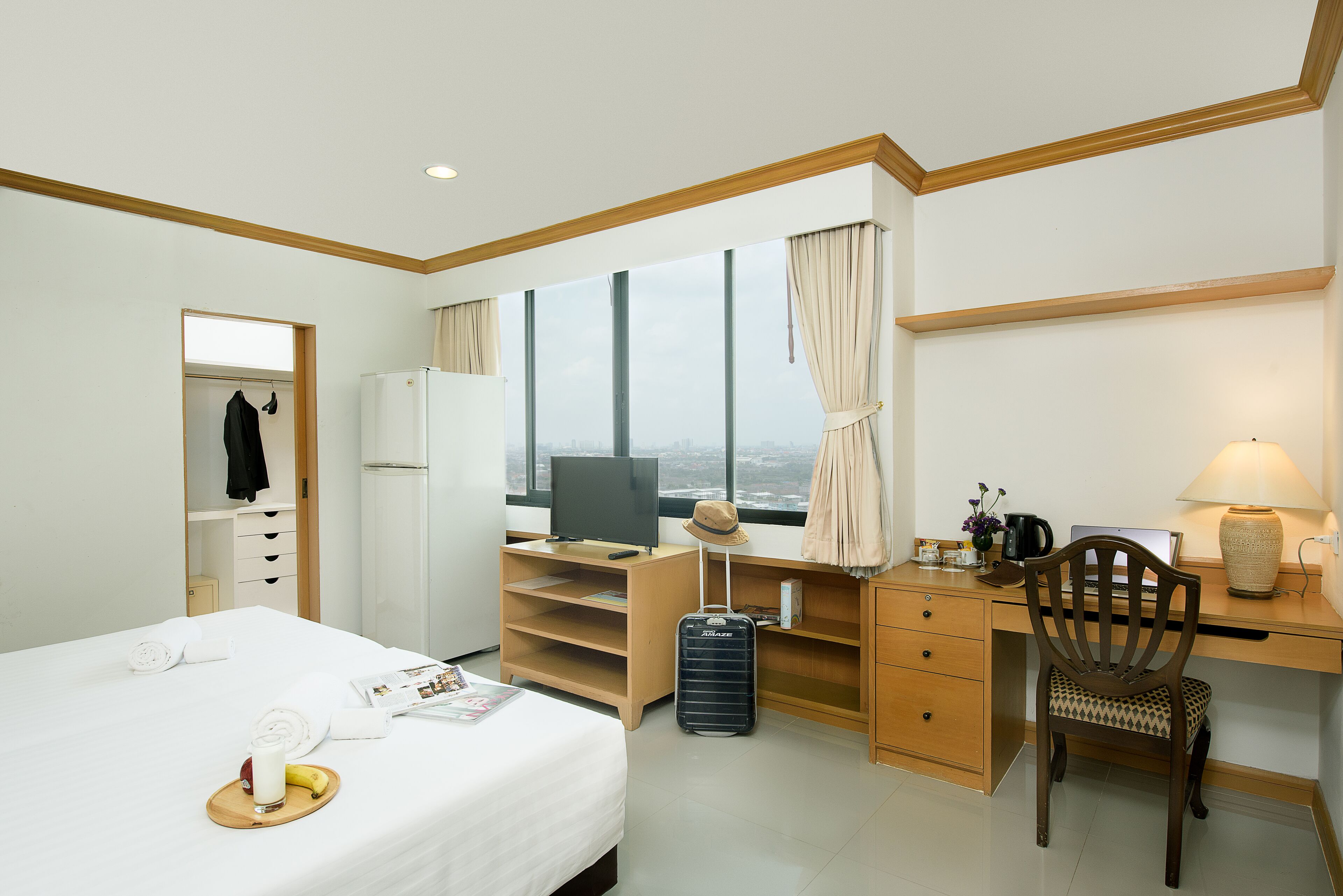 A hotel room with a bed, a desk with a computer, a chair, a suitcase, and a window with curtains.