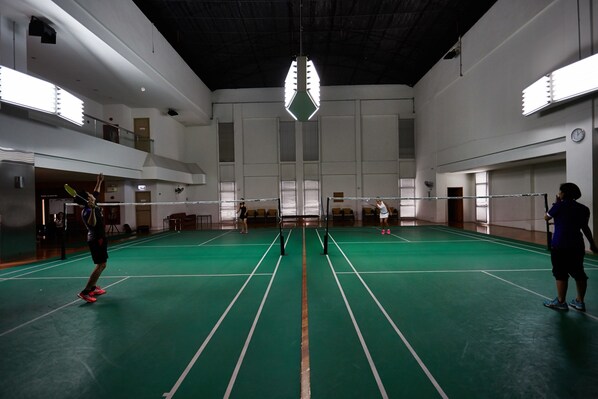 An indoor badminton court with a high ceiling, multiple windows, and a clock on the wall.