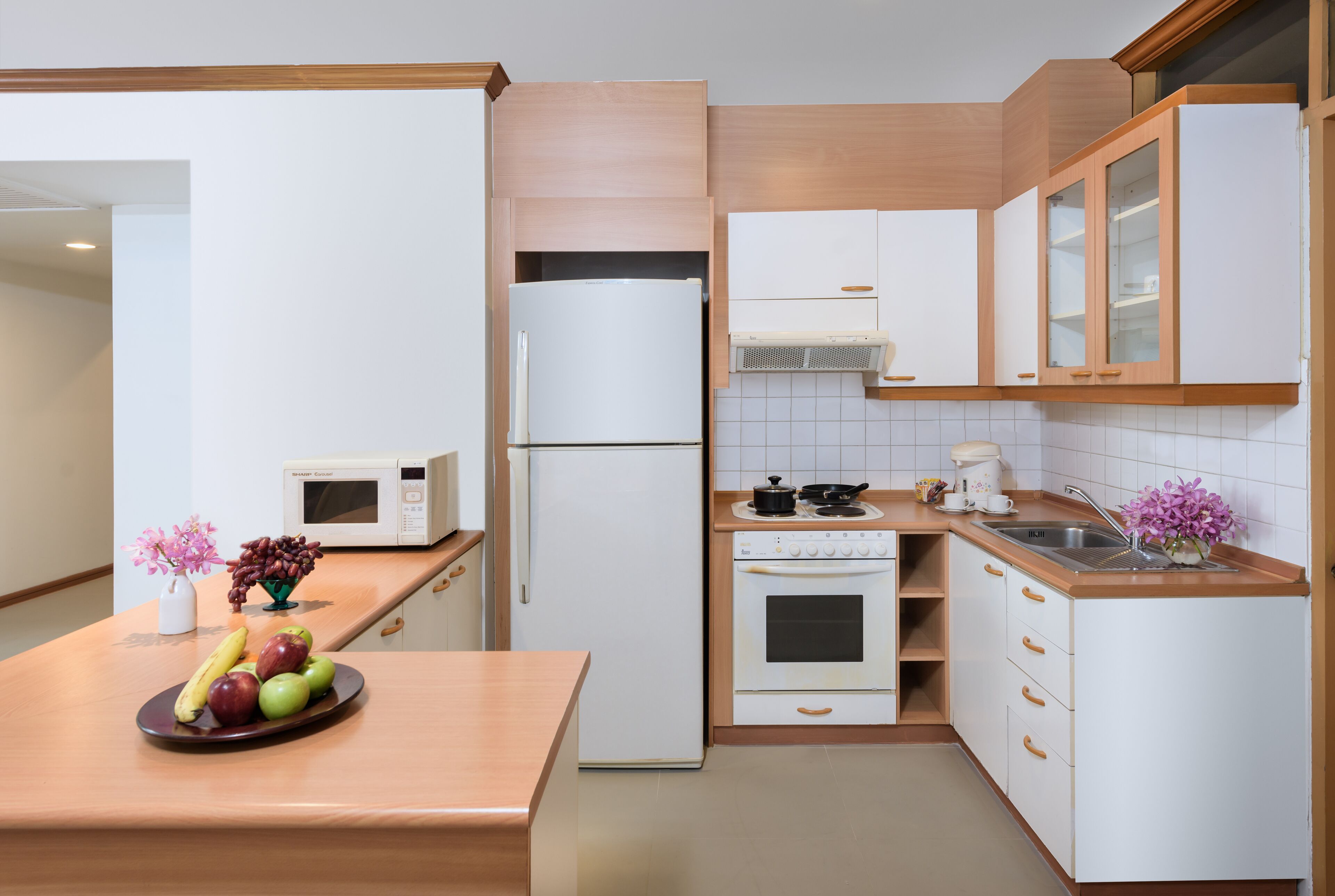 Een moderne keuken met witte kastjes, een koelkast, magnetron, oven en spoelbak. Op het aanrecht staat een fruitschaal en een vaas met bloemen.