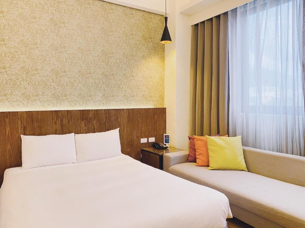A hotel room with a large bed, a sofa, a wooden headboard, a nightstand, and a window with curtains.