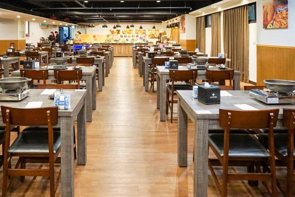 A restaurant with multiple tables and chairs, a counter with a display of food, and a kitchen area in the background.