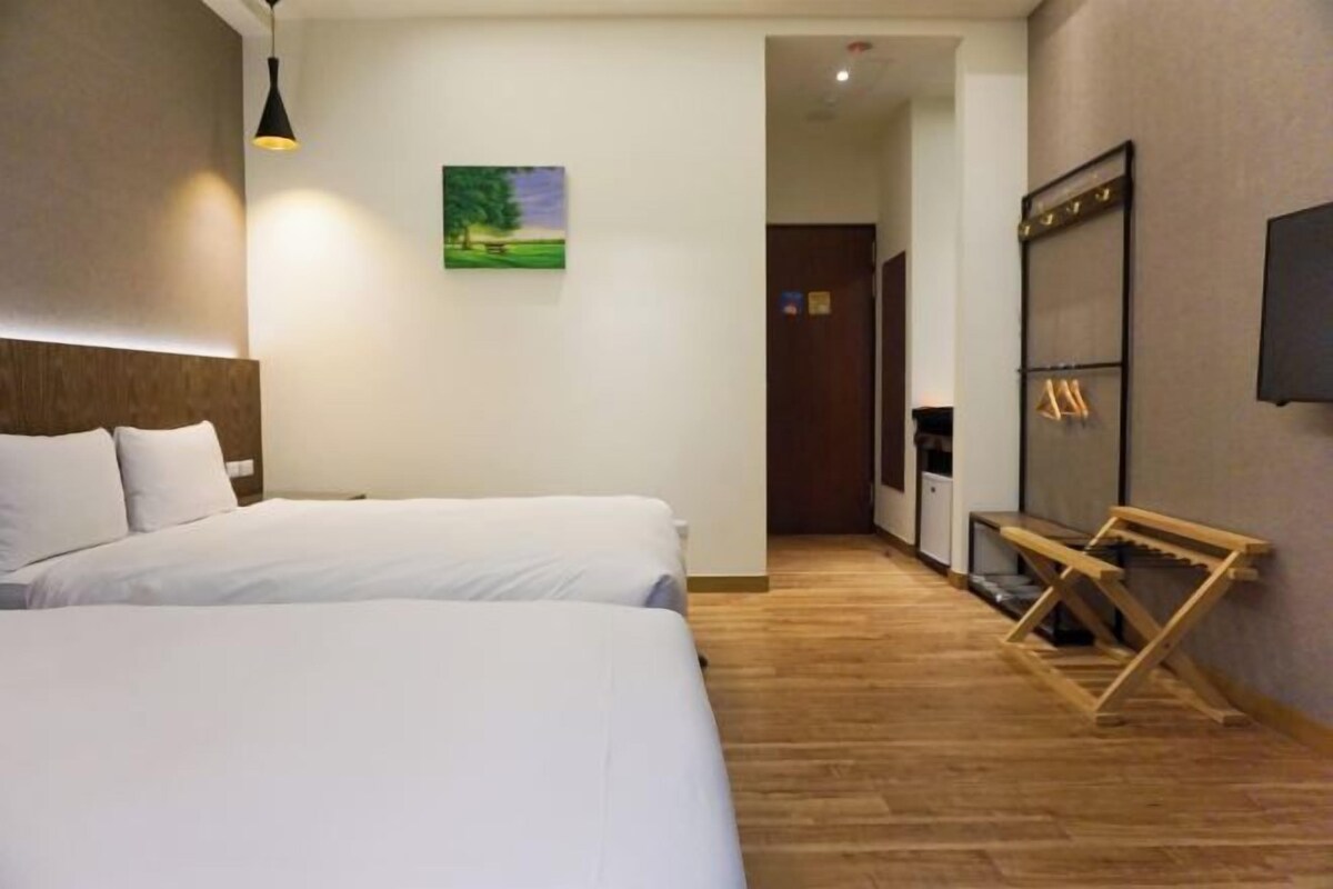 A hotel room with two beds, a wooden floor, a painting on the wall, and a TV mounted on the wall.