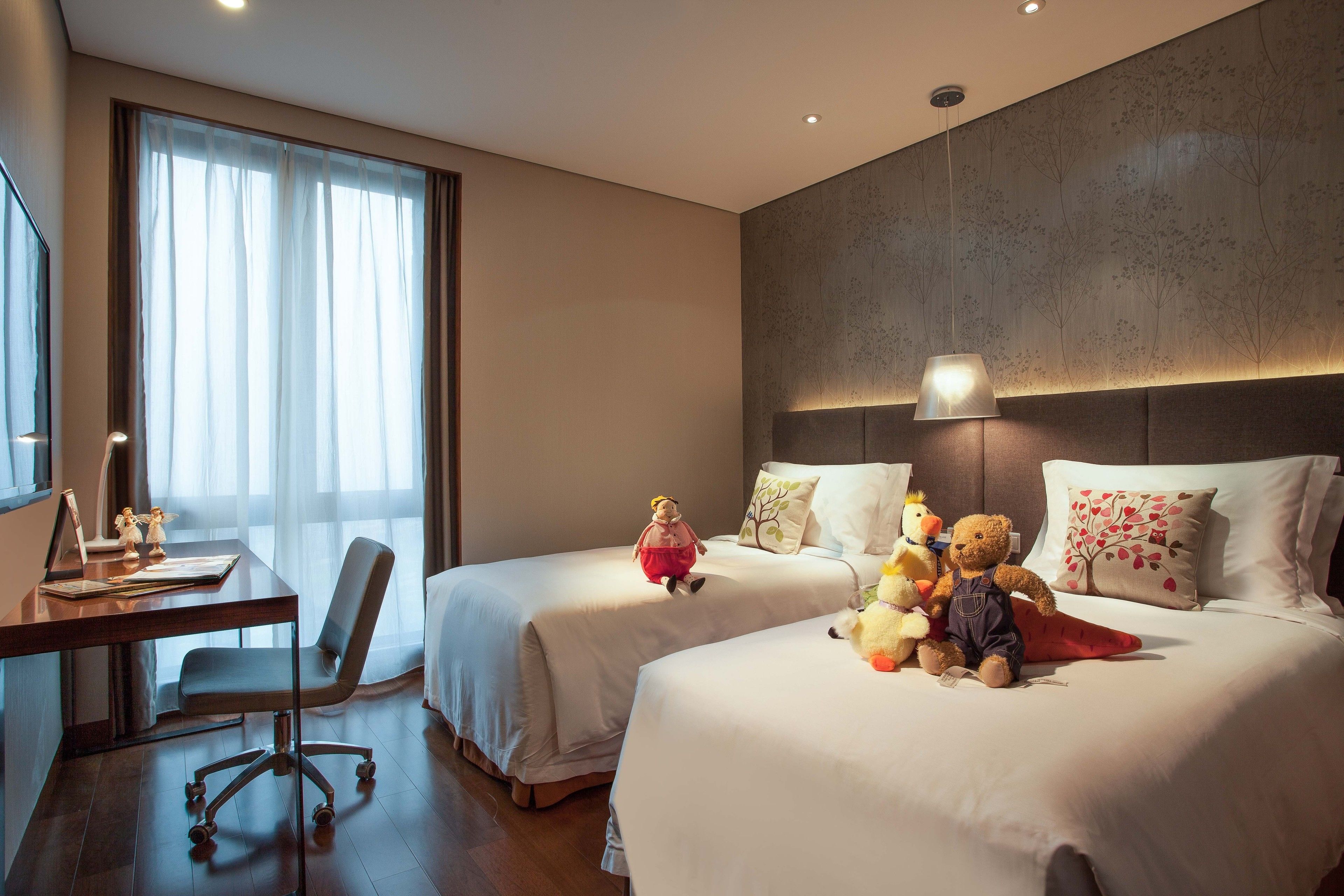 A hotel room with two beds, a desk, and a chair. There are decorative pillows and stuffed animals on the beds.