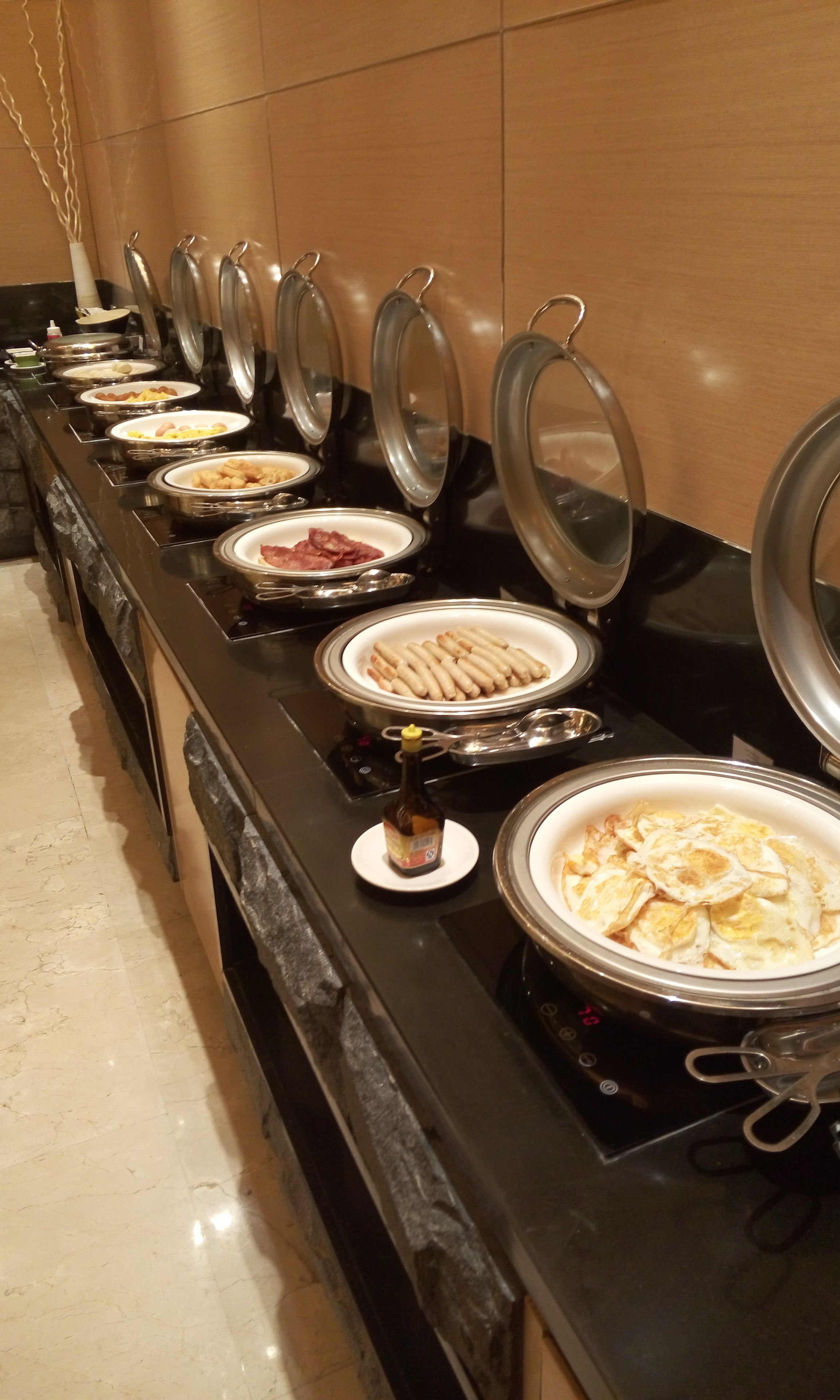A buffet line with various food items, including sausages, eggs, and meats, arranged on a counter with lids.