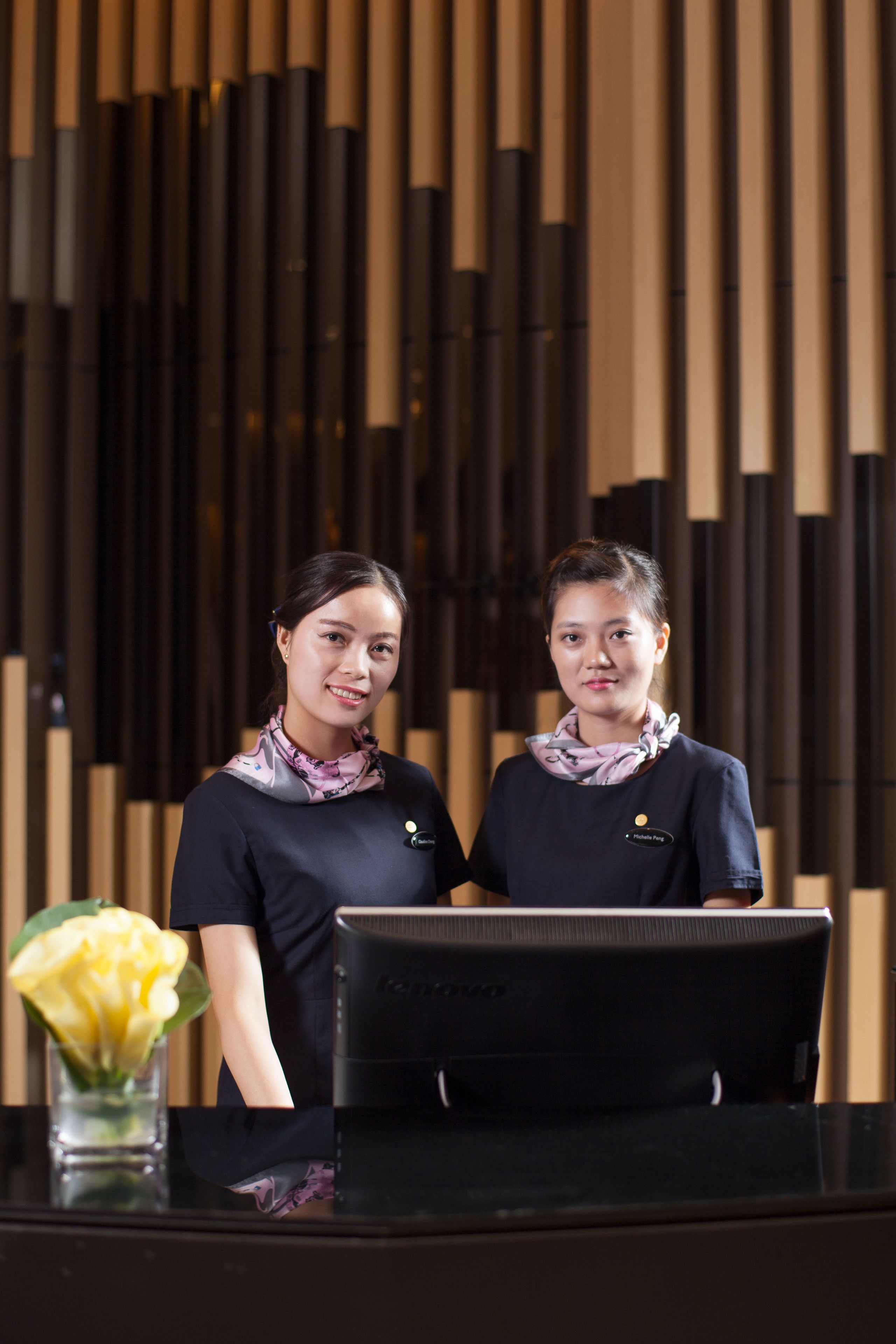 Two individuals standing at a reception desk with a modern design, featuring vertical wooden panels in the background.