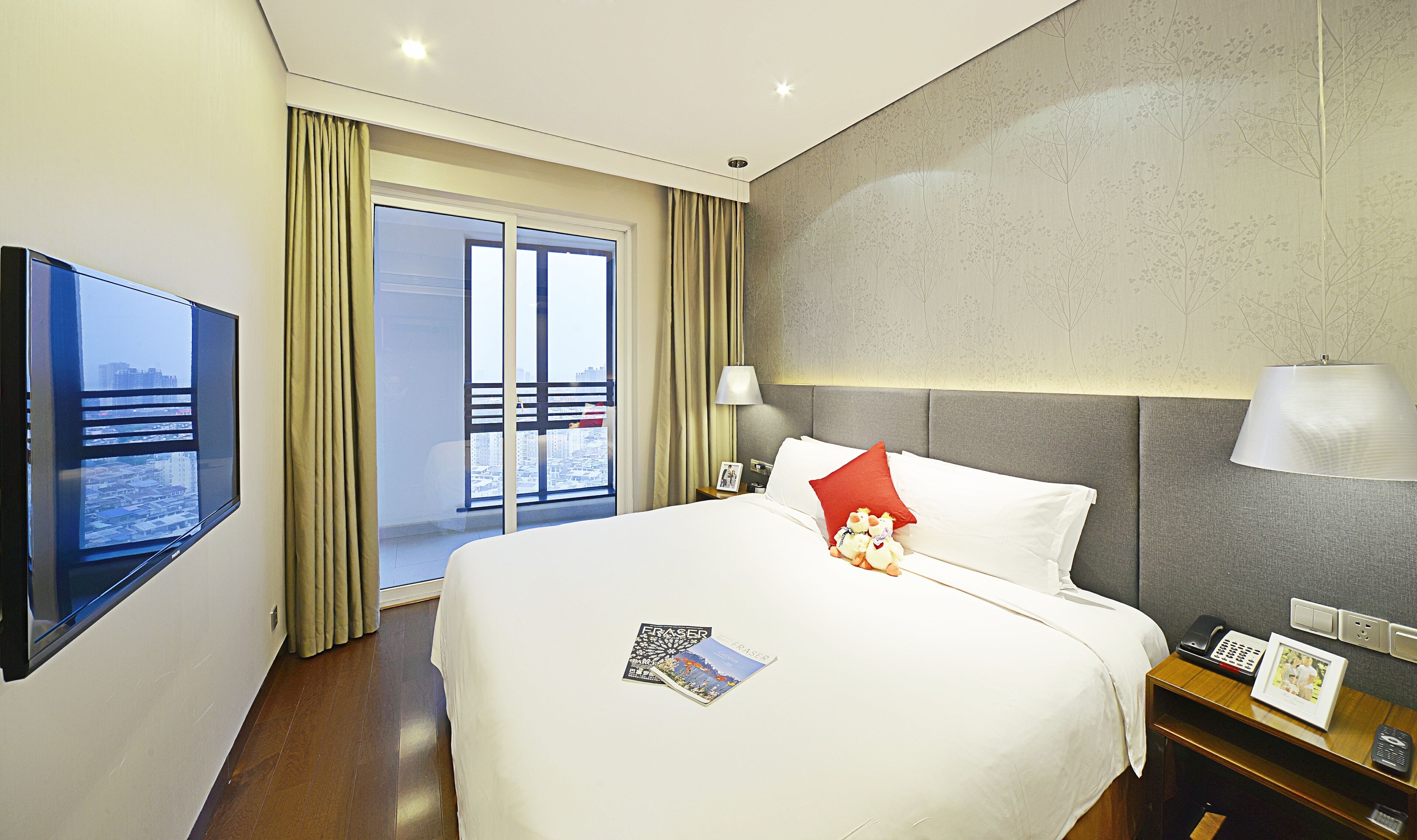 A hotel room with a bed, a TV, a bedside table with a lamp, a phone, and a picture frame.