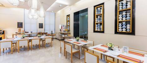 A modern dining area with multiple tables and chairs, decorative lighting, and wall-mounted shelves with dishes.