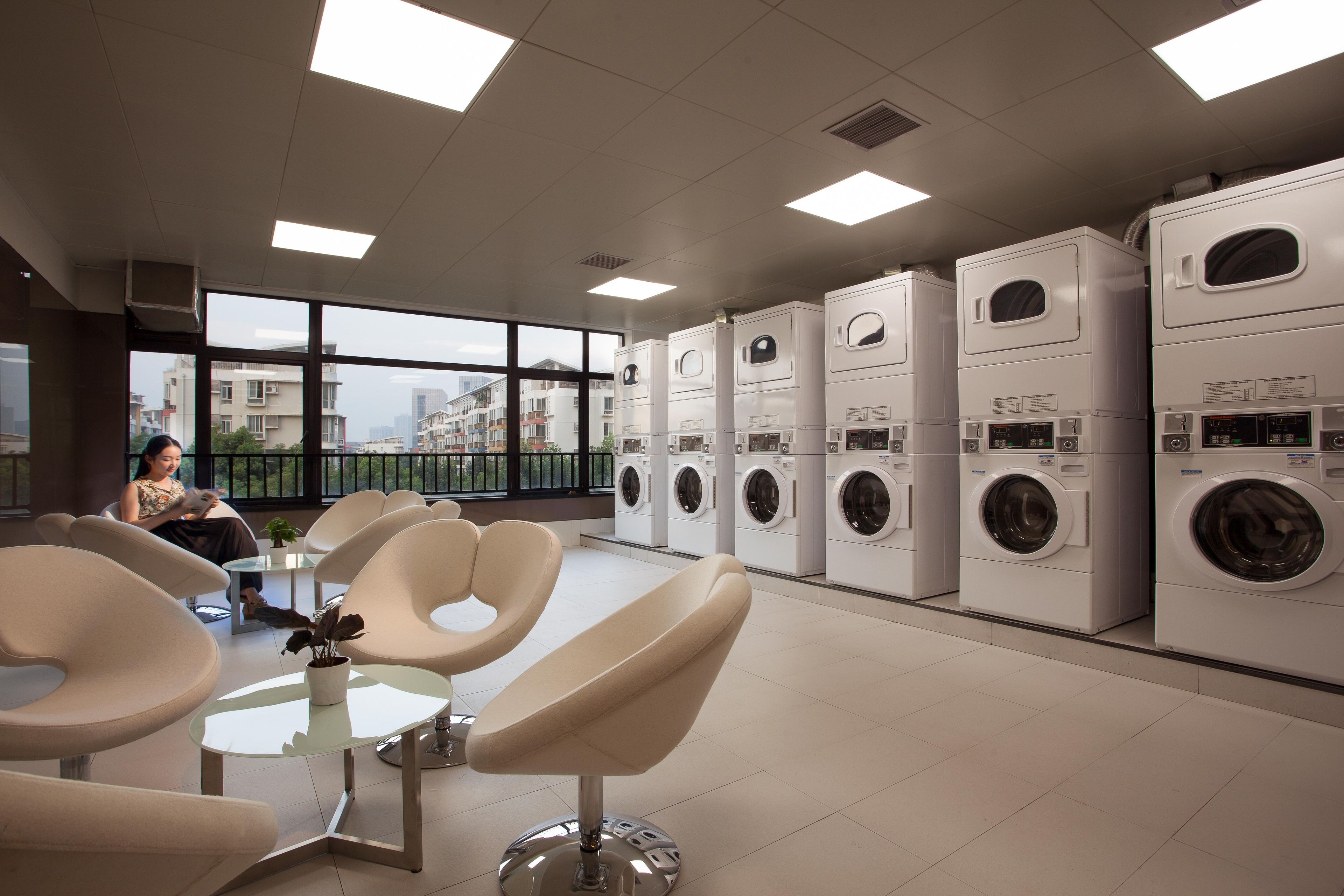 A laundry facility with multiple washing machines, seating areas, and large windows offering a view of the city.