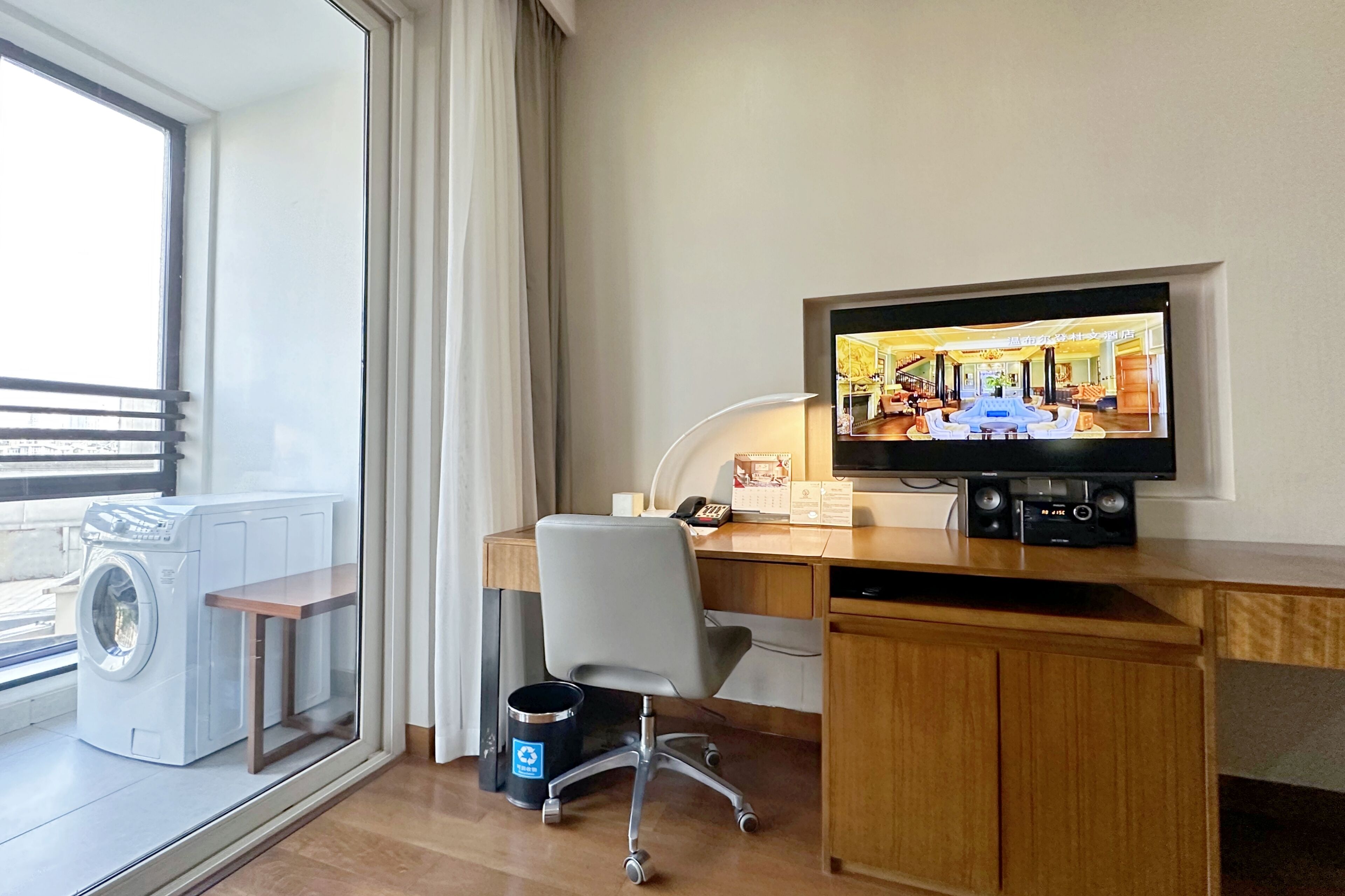 A hotel room with a desk, chair, washing machine, and a television displaying a hotel interior.