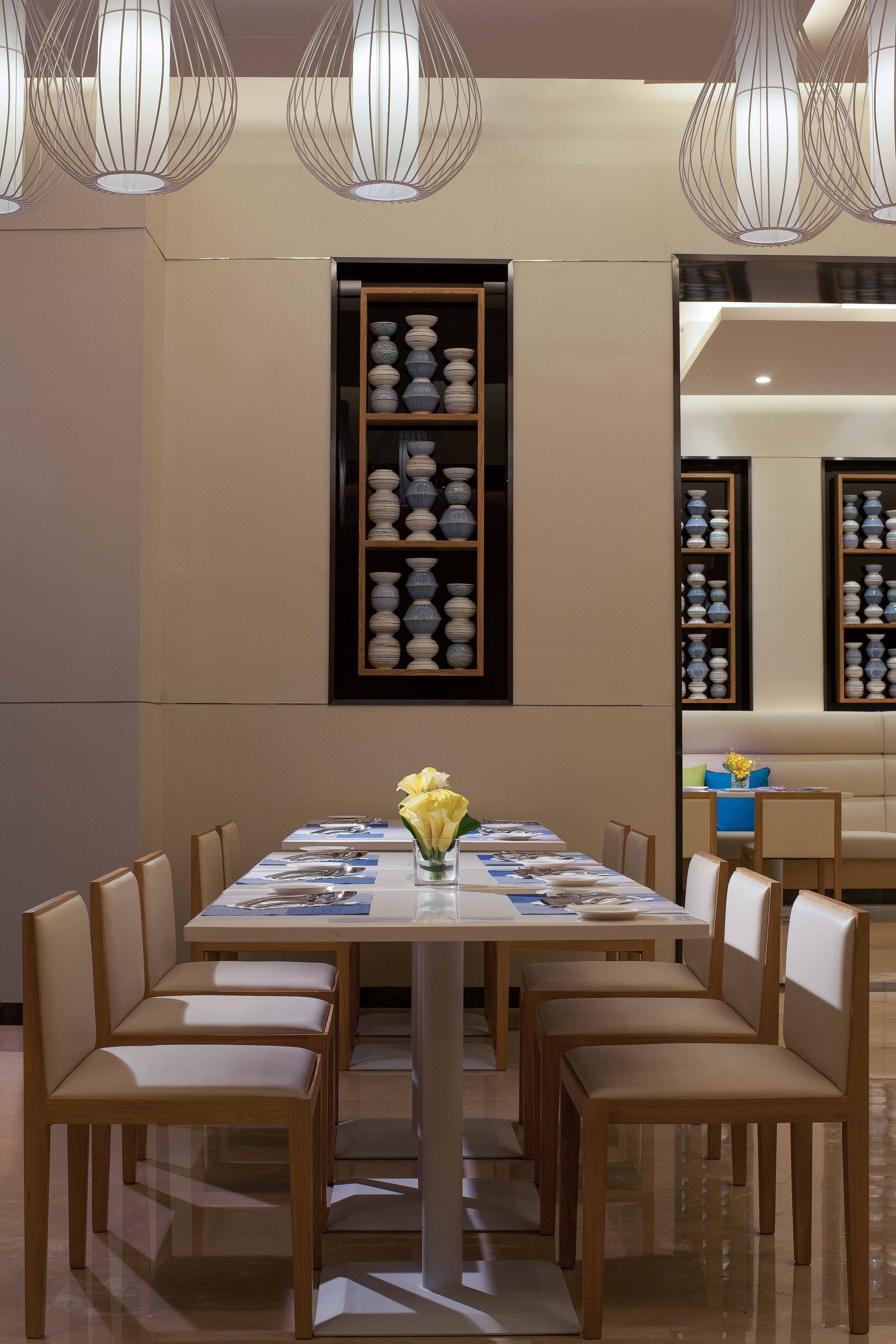 A dining area with a long table, multiple chairs, and a central vase with yellow flowers. Shelves with white and blue bowls are mounted on the wall.