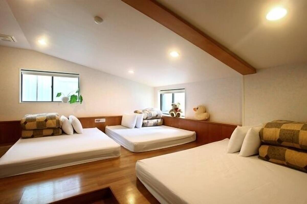 A room with three beds arranged in a U-shape, wooden flooring, and a sloped ceiling with recessed lighting.