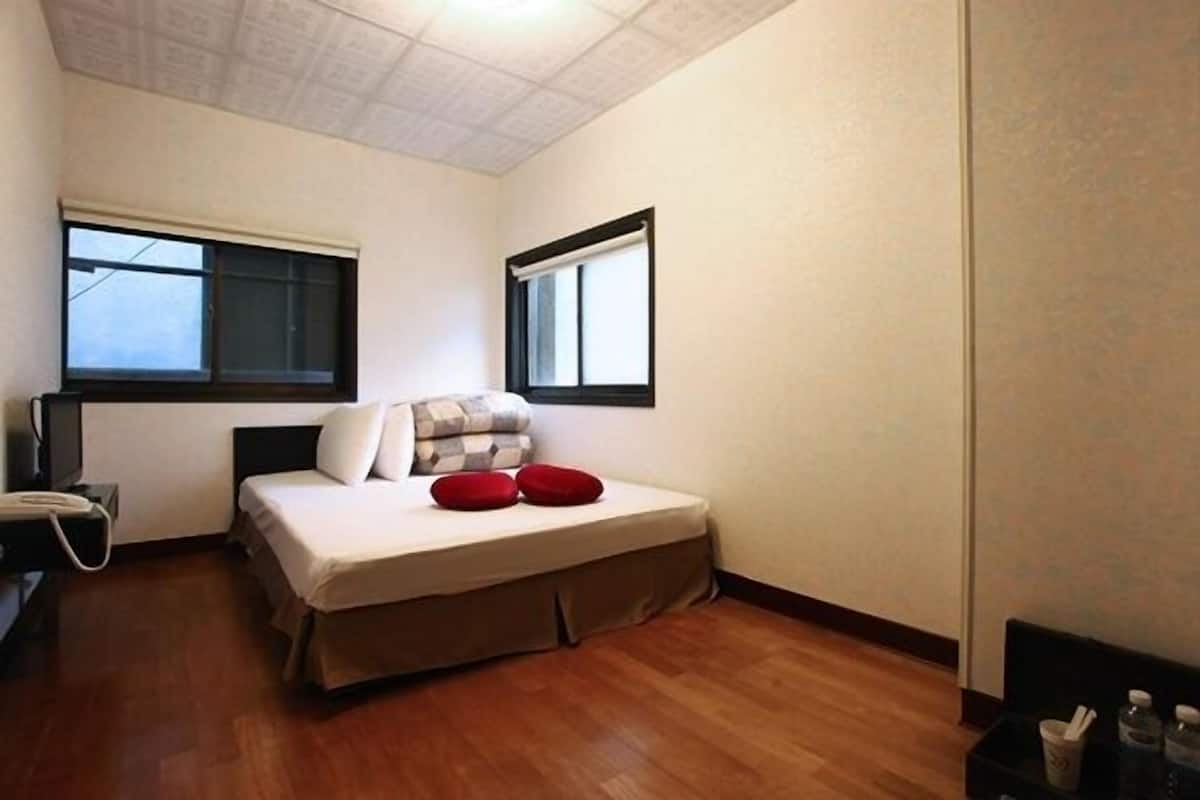 A small, clean room with a bed, two windows, a bedside table, and a telephone.