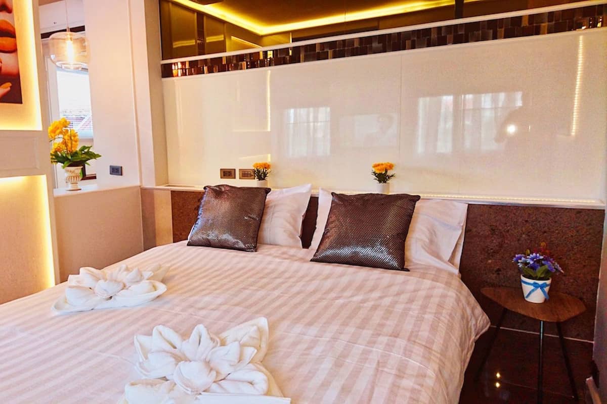 Habitación de hotel con una cama grande, dos almohadas, una mesita de noche, un florero con flores y un cuadro colgado en la pared.