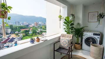 A balcony with a view of a cityscape, a washing machine, a chair, and various potted plants.