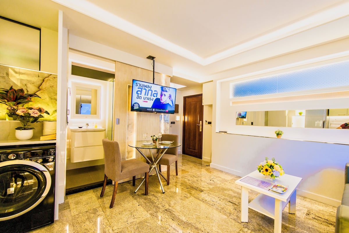 Una habitación de hotel moderna con piso de mármol, un área de comedor con una mesa de vidrio, un televisor montado en la pared y una pequeña mesa con flores.