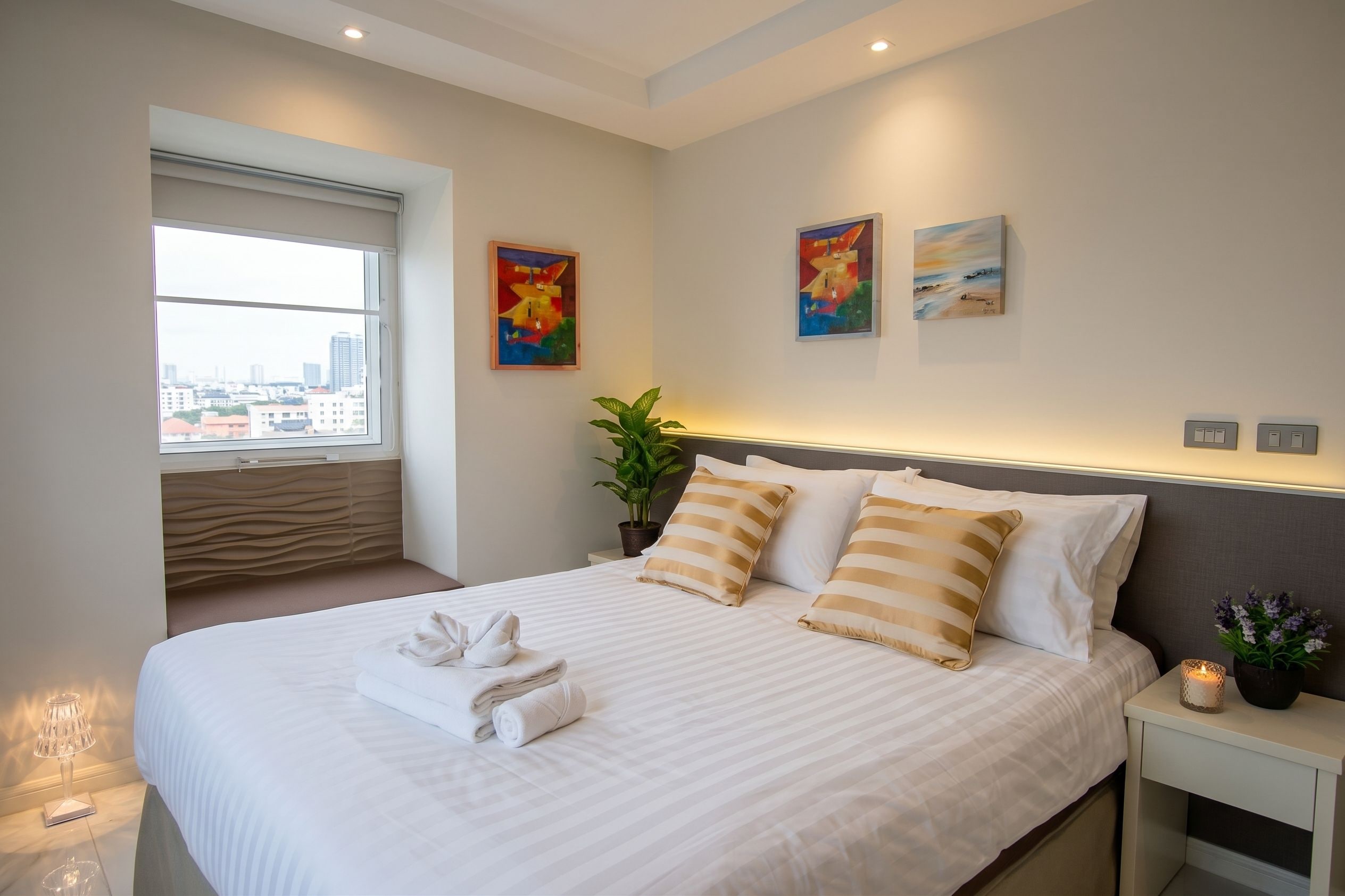 A hotel room with a large bed, two paintings on the wall, a window with a view of buildings, and a bedside table with a lamp and a vase of flowers.
