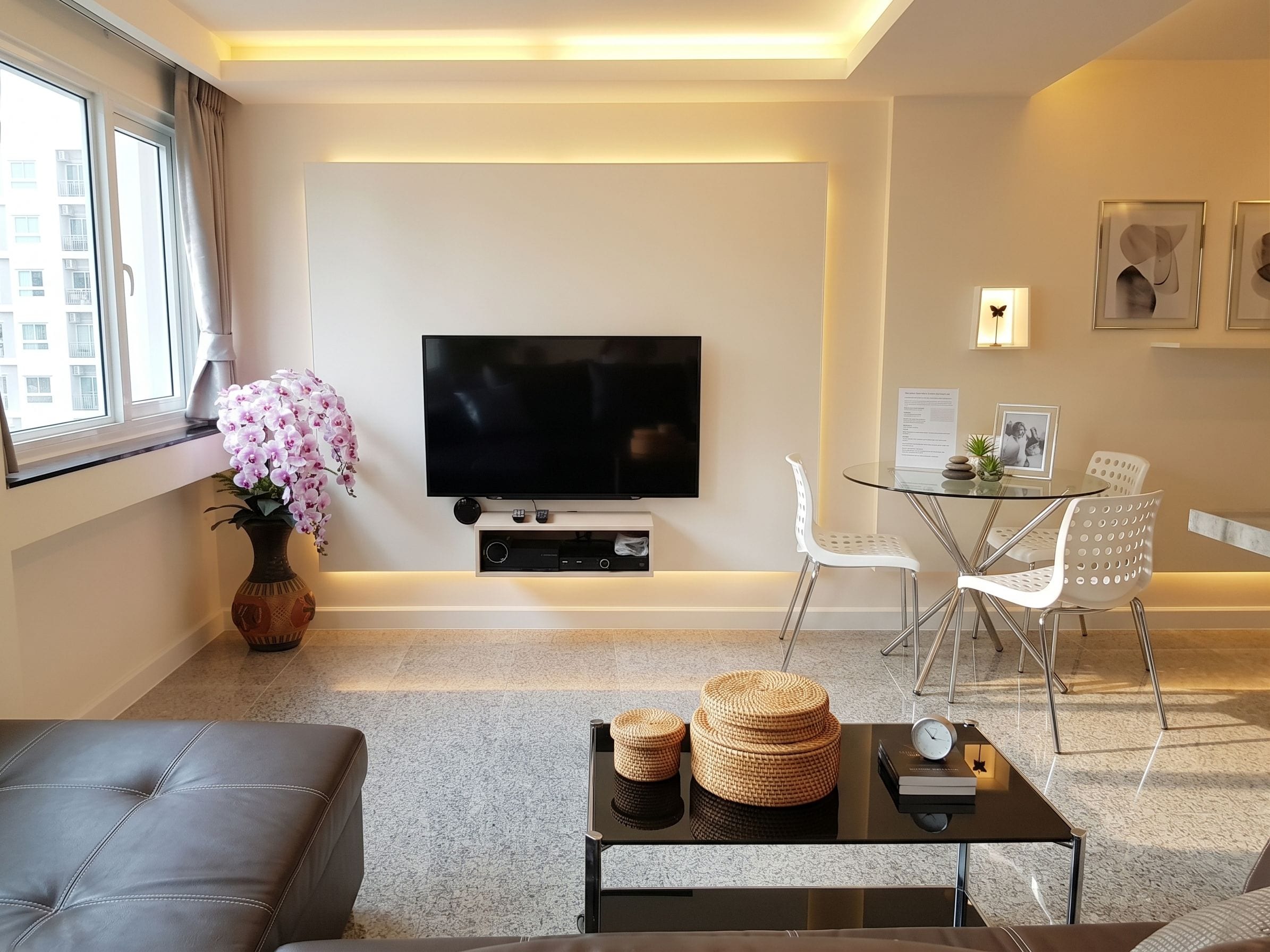 A modern living room with a flat-screen TV, a sofa, a coffee table, a dining area with a glass table and chairs, and a vase with flowers.