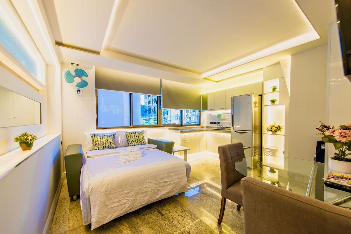 Una habitación de hotel moderna con una cama, una mesa de vidrio, una silla, una kitchenette y vistas a la ciudad.