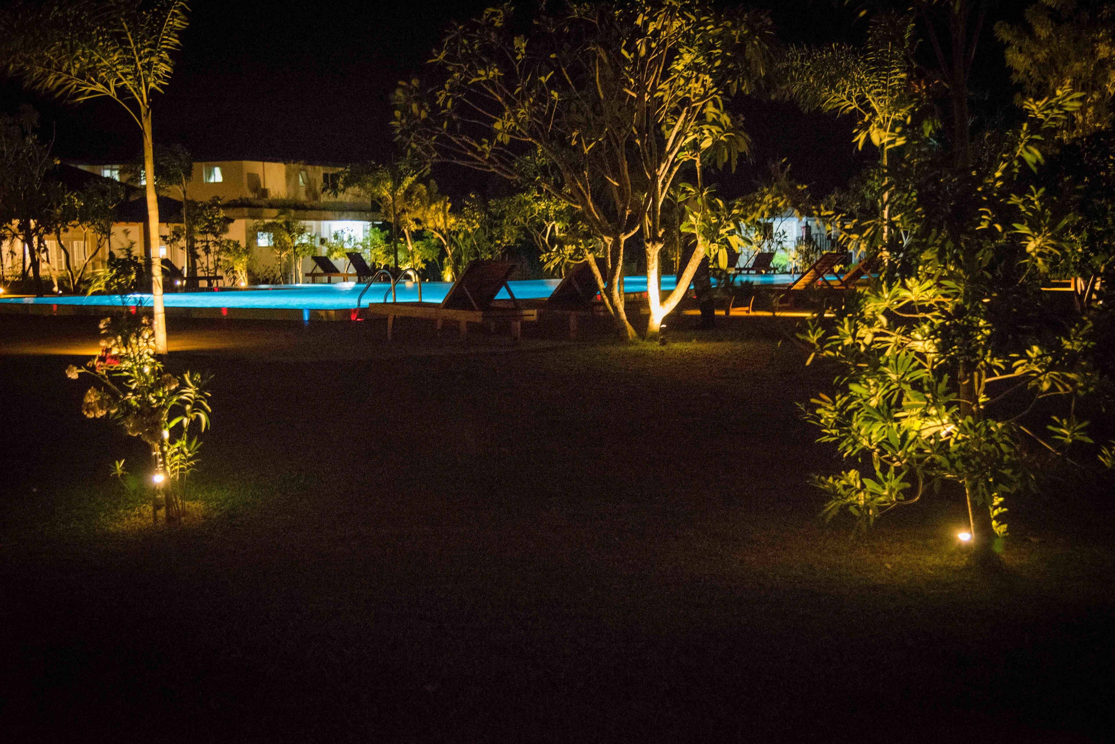 A nighttime view of a resort with a swimming pool, lounge chairs, and trees.