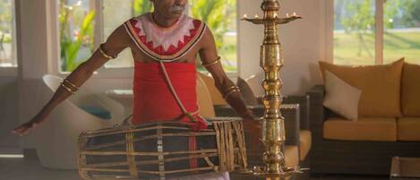 Een persoon in traditionele kleding die drum speelt in een kamer met een grote messing lamp, een bank en een raam met jaloezieën.