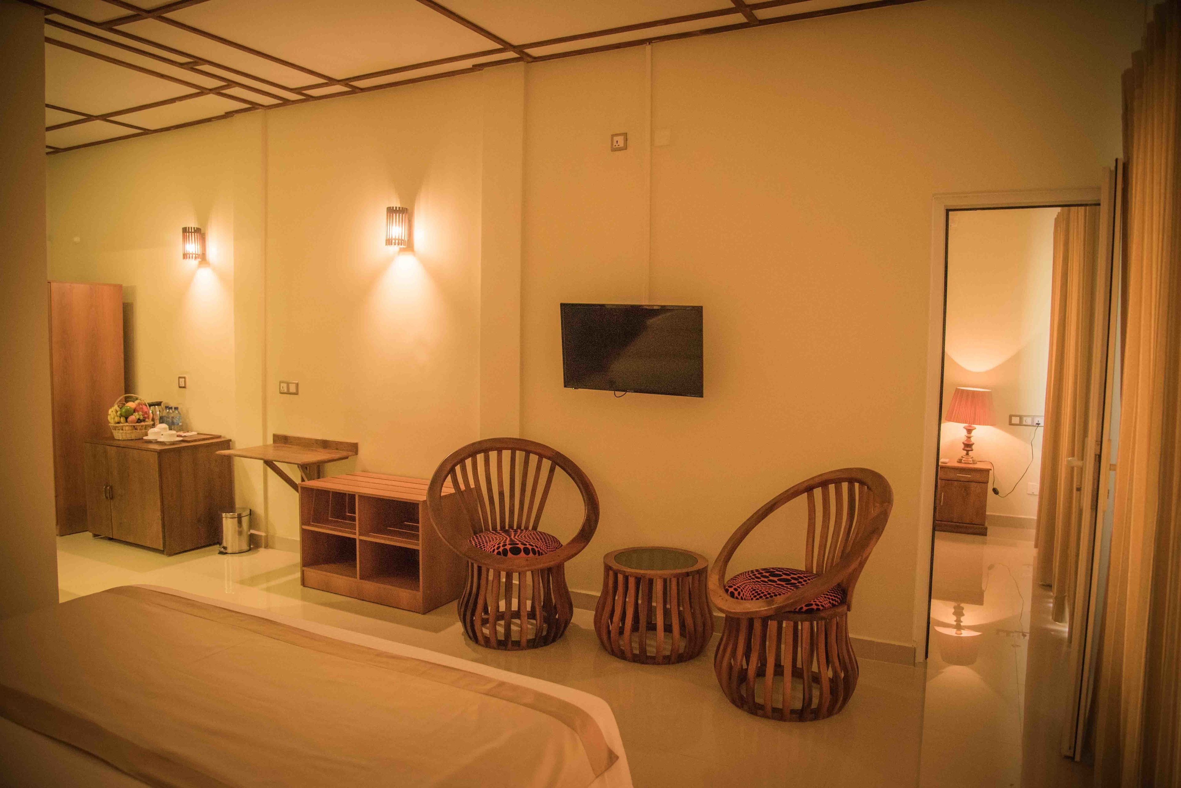 A hotel room with a bed, two wooden chairs, a small table, a TV mounted on the wall, and a cabinet with a basket of fruit.