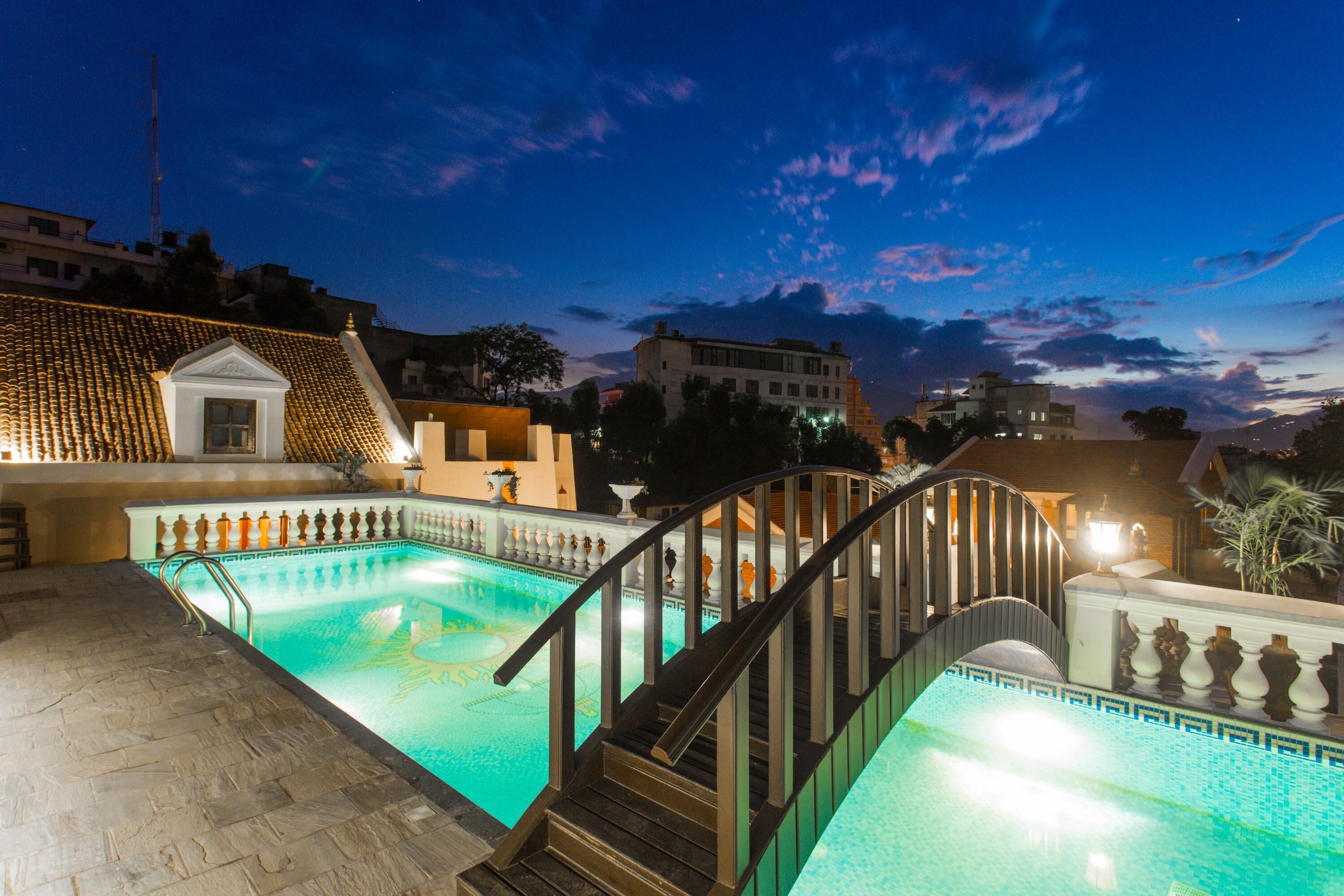 A swimming pool with a wooden staircase leading up to a balcony, surrounded by buildings and a clear sky.