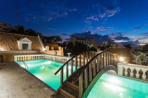 A swimming pool with a wooden staircase leading up to a balcony, surrounded by buildings and a clear sky.