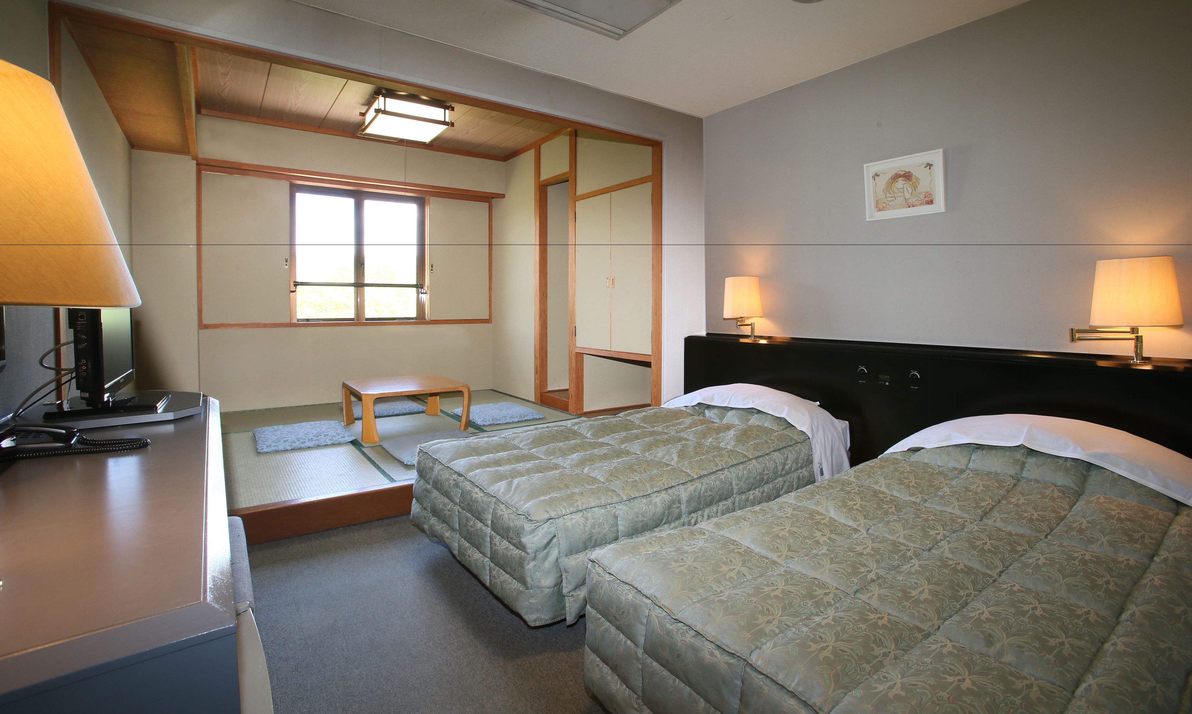 A hotel room with two beds, a desk with a lamp, a small table, and a window with natural light.