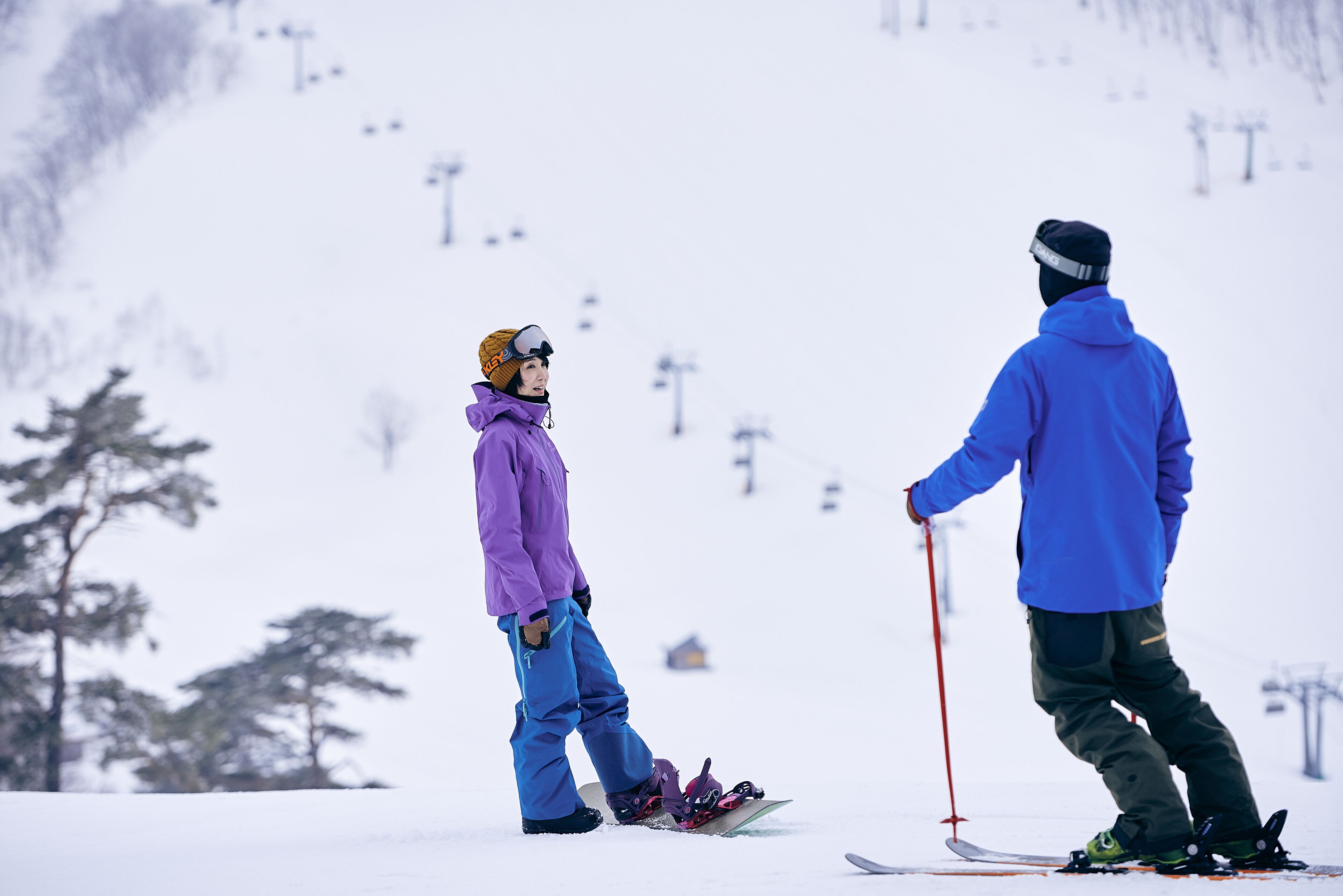 A person in a purple jacket and blue pants standing on a ski slope with a ski lift in the background.